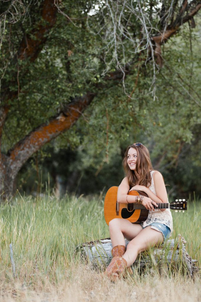 Creative senior photo of a girl wearing cowboy boots and shorts in Verdi, while she rests her hand on her acoustic guitar by Reno Senior Photographer.
