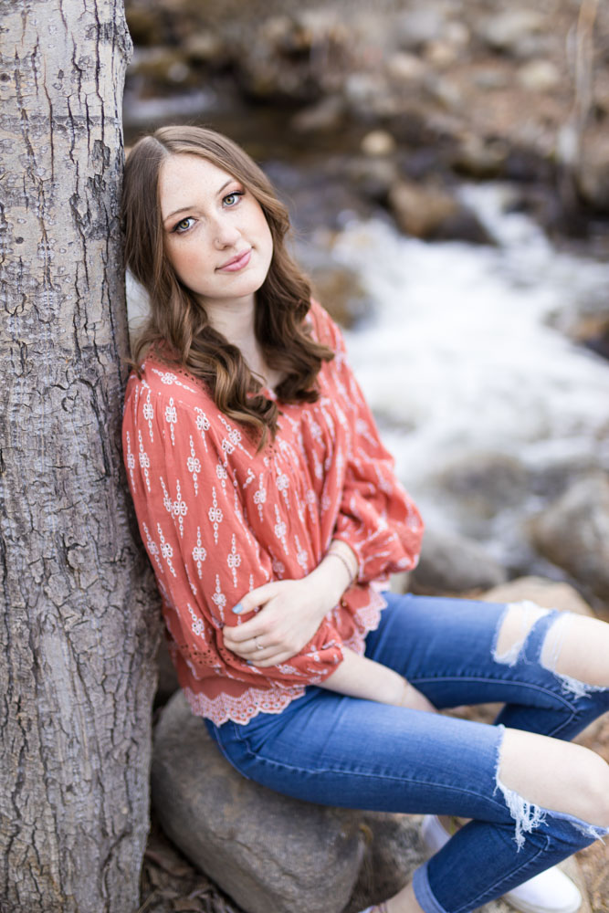 A female student wearing coral in boho chic style sits on a rock in front of a raging creek in the spring for her reno high school senior photography experience with Fifth and Chestnut