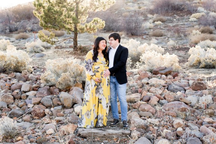 Pregnant woman in yellow and gray floral dress stands with her husband among desert foliage for a maternity photo shoot in Reno by Reno maternity photographer