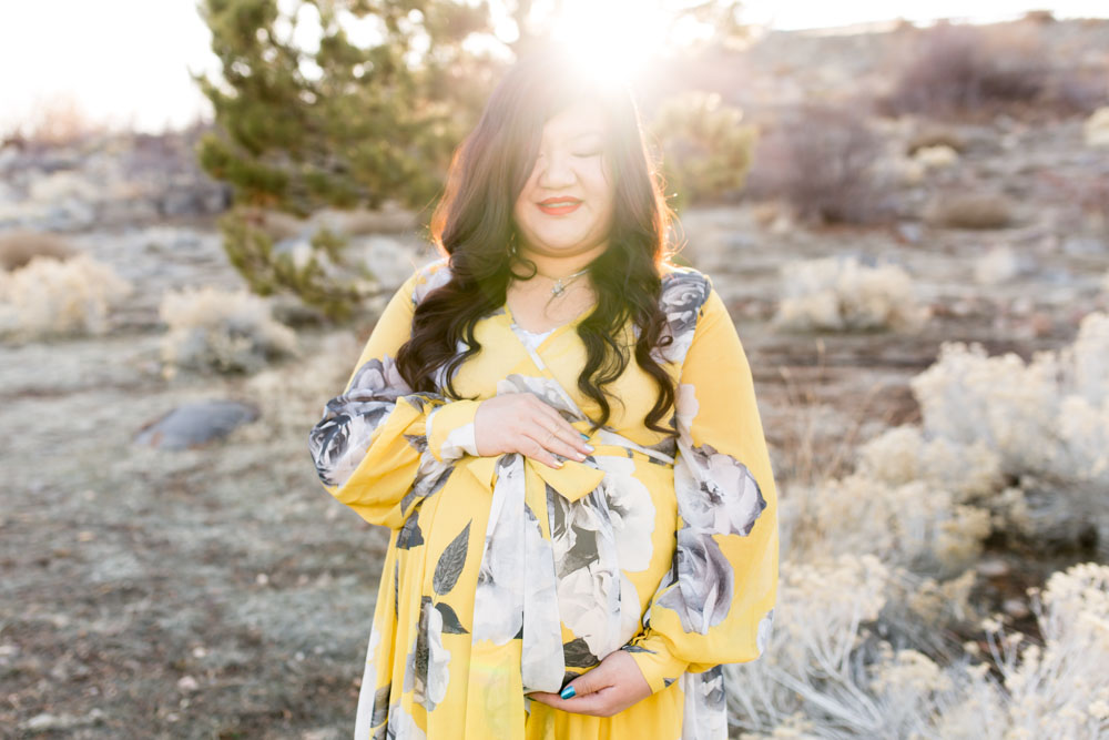 Pregnant woman in yellow and gray floral dress holds her pregnant belly and looks down as the sun peeks over her the top of her head in Reno, NV