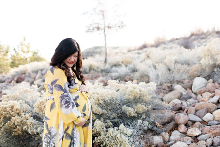 Pregnant woman in yellow and gray floral dress holds pregnant tummy while looking down surrounded by high desert foliage in Reno, NV for maternity portraits by Reno Maternity Photographer