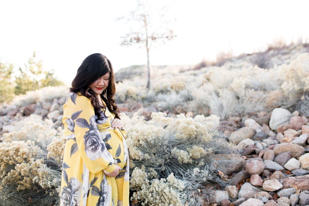 Pregnant woman in yellow and gray floral dress holds pregnant tummy while looking down surrounded by high desert foliage in Reno, NV for maternity portraits by Reno Maternity Photographer