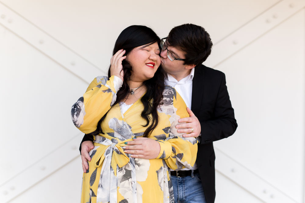 Husband kisses his pregnant wife on the cheek in yellow and gray floral dress in front of a white barn door in Reno, NV