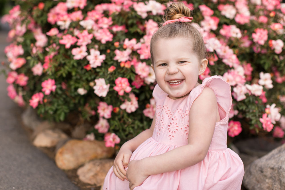 A three year old blonde girl wearing a pink dress smiles in a park filled with lovely foliage for her three year Reno Child Portraits by Reno Child Photographer