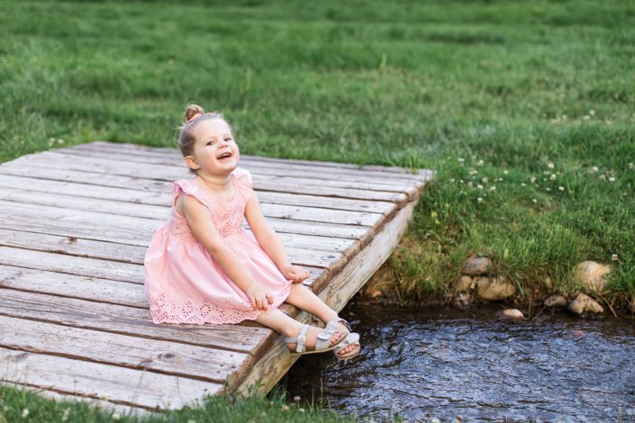 A three year old blonde girl wearing a pink dress smiles in a park filled with lovely foliage for her three year Reno Child Portraits by Reno Child Photographer