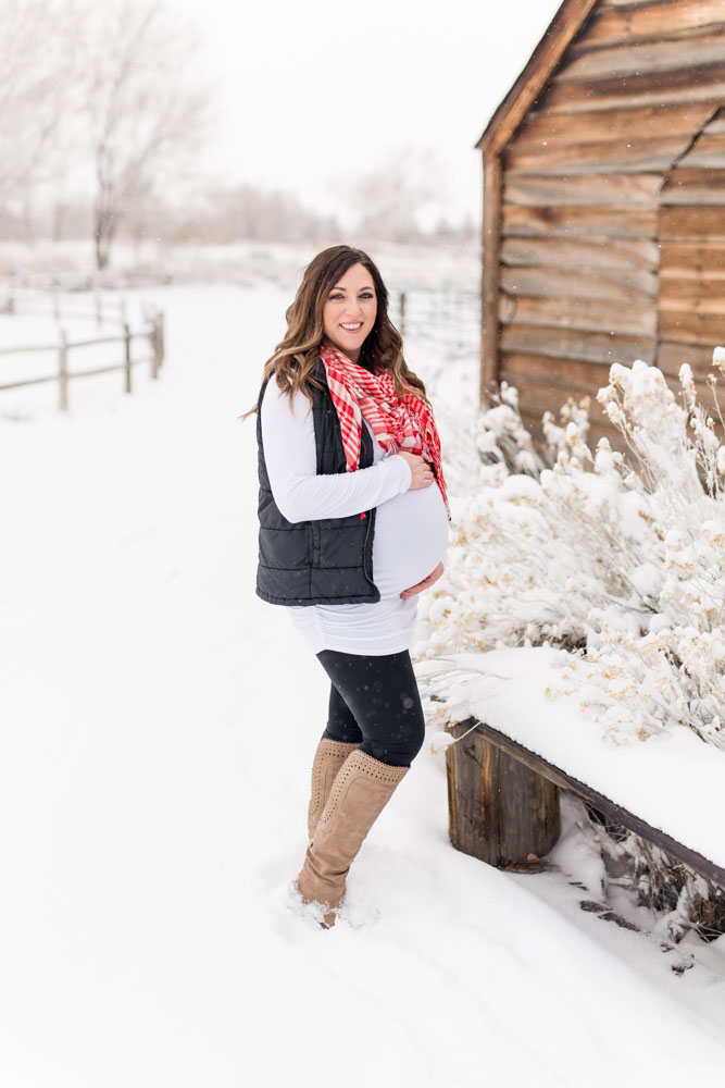 Pregnant woman wearing red and white plaid scarf and a black vest over a white shirt holds her baby belly in front of a wooden bench and wooden building at Bartley Ranch in Reno for her snowy Reno maternity photos by Reno maternity photographer