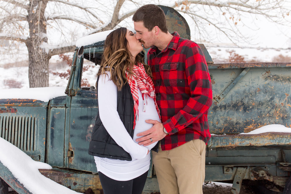 Pregnant couple kisses in front of rusty old truck at Bartley Ranch in Reno for their snowy Reno maternity photos by Reno maternity photographer