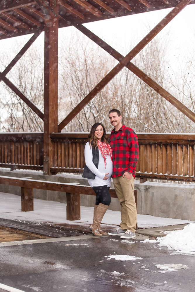 Couple stands under covered bridge while it snows heavily around them at Bartley Ranch in Reno for their snowy Reno maternity photos by Reno maternity photographer