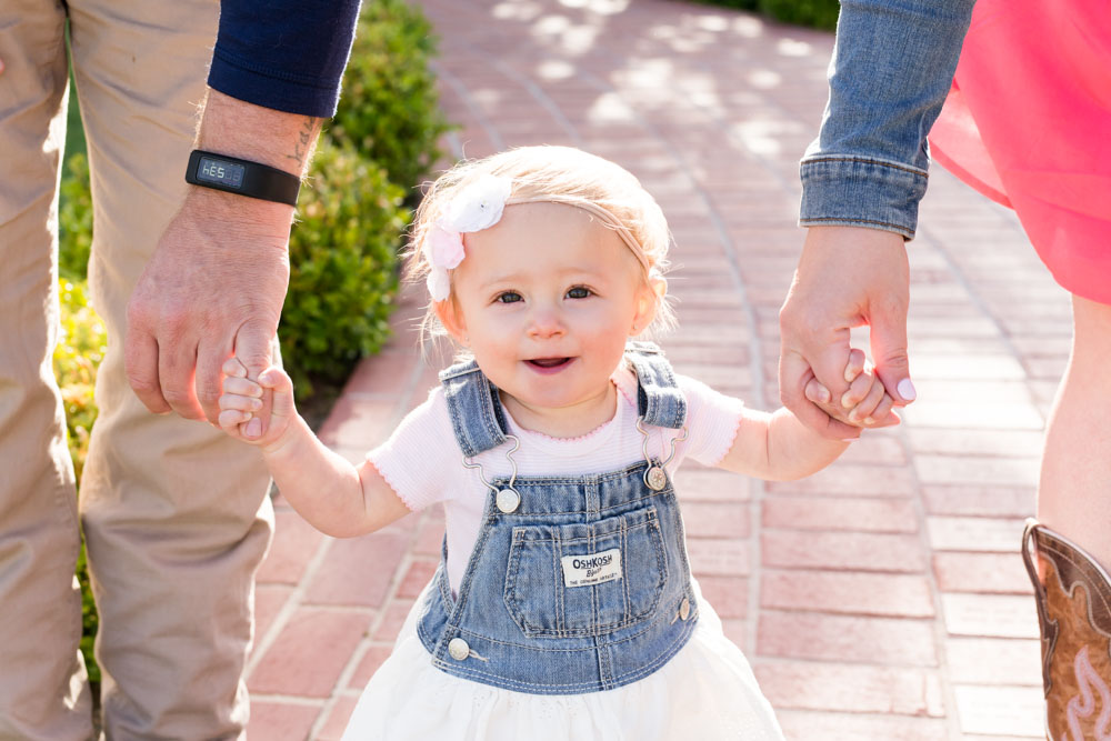 one year old baby girl in osh kosh overall dress with lace and denim holds her mom and dad's hands walking down a brick path at bartley ranch for her reno one year photos by Reno Child Photographer
