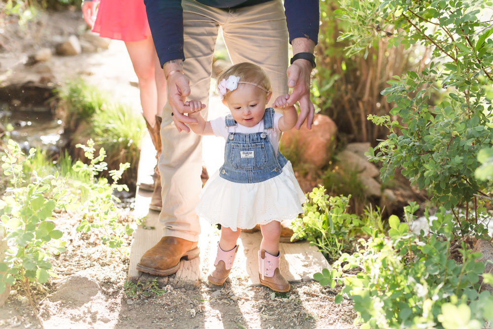one year old baby girl wearing a dress walking down a dirt path holding onto her dad's hands for one year photos in reno by reno child photographer