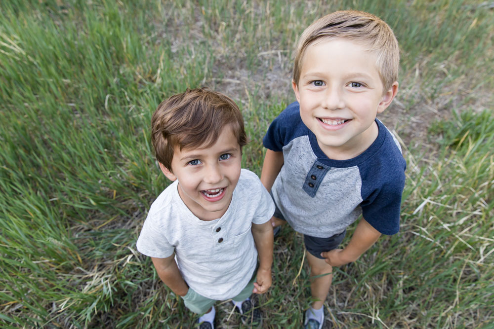 Two boys laugh looking upward into the camera as they stand in a beautiful summer meadow filled with grass