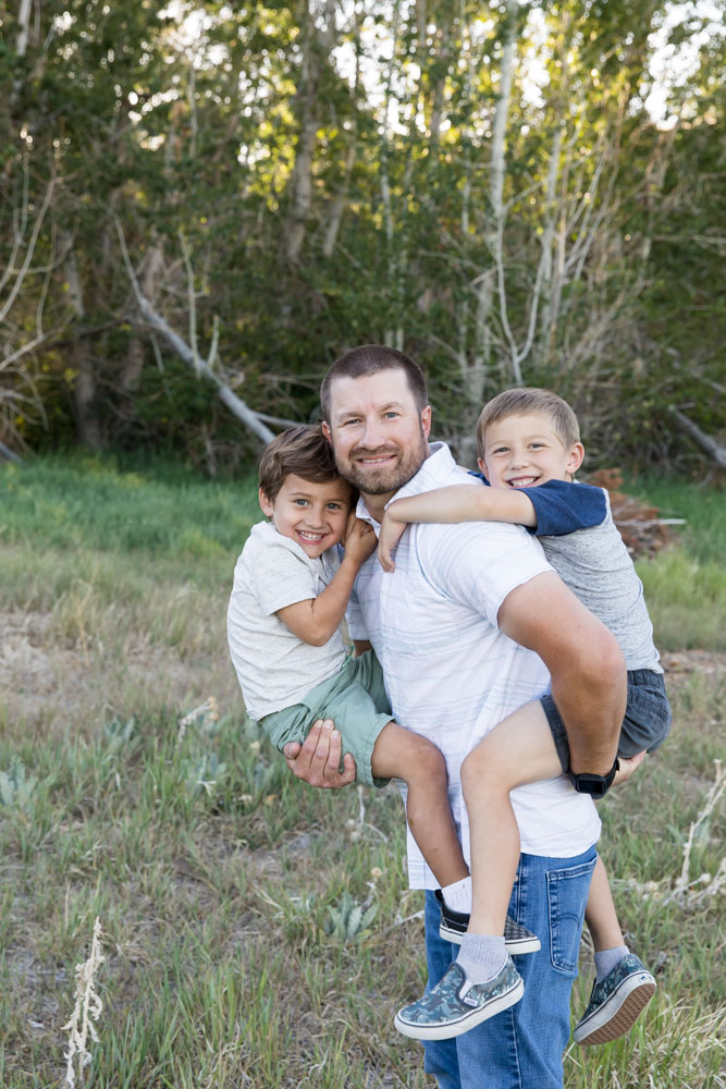 A dad carries his two little boys on his back and chest as they play in a lush summer meadow for Reno Family Portraits by Reno Family Photographer