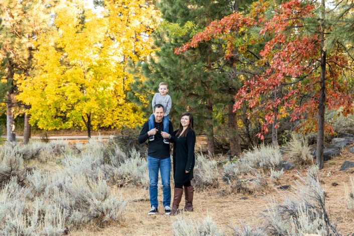 A boy perches on top of his dad's shoulders for a family photo in front of red and yellow trees at Callahan park in Reno, NV