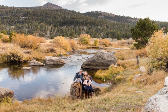 A family of four wearing fall clothing sits on a big rock near a river for their Hope Valley Family Portraits by Hope Valley Family Photographer with towering mountains behind them