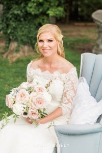 A gorgeous blonde bride smiles at the camera holding a whimsical wedding bouquet on a country french blue chair at a private estate