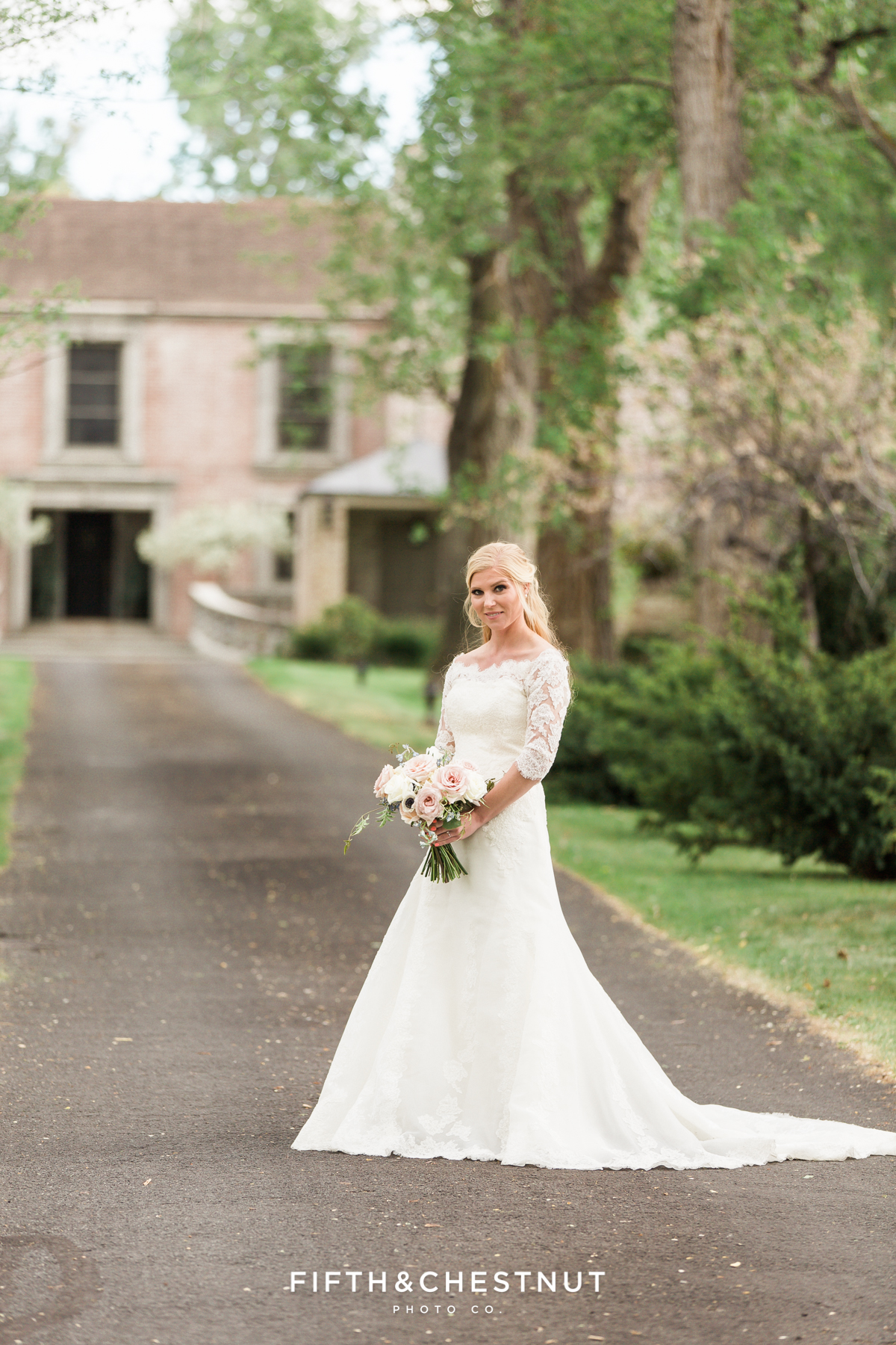 Happy bride in lace dress standing in an oak-lined pathway leading to a stone and brick private estate