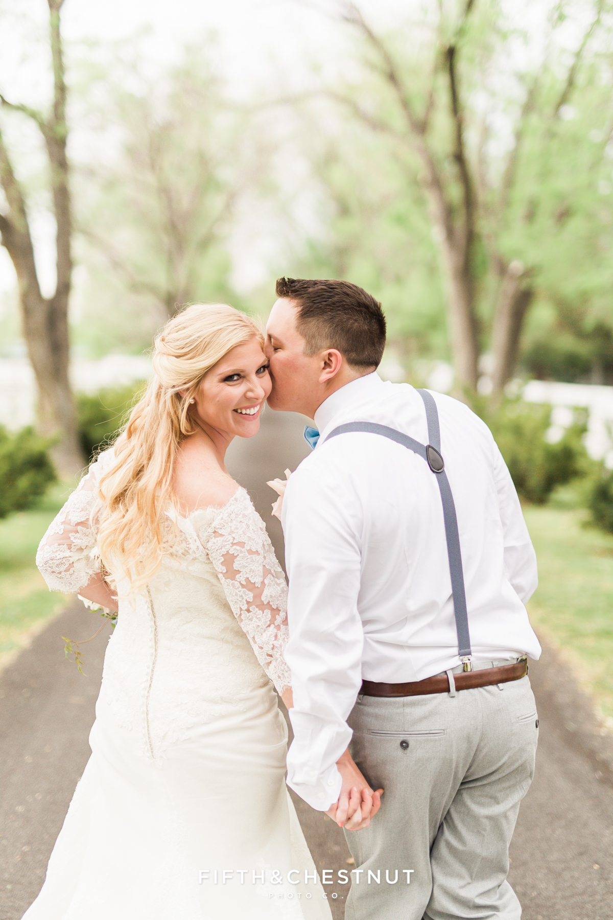 Bride laughing as her groom kisses her cheek as they walk down an oak-lined path