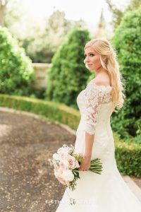 Bride looks into the distance while standing in front of hedges and brick wall
