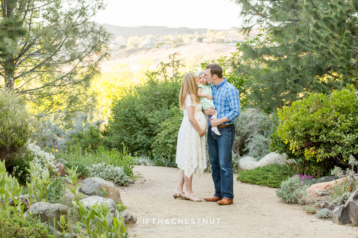 Mom and dad kiss their daughter's cheeks while standing in a path lined with flowers and plants at Rancho San Rafael park in reno