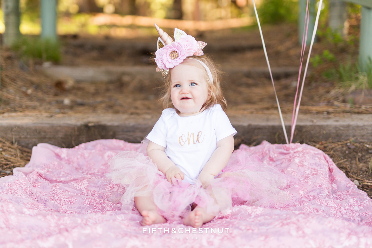 One year old girl sitting on floral pink blanket wearing a "One" onsie and unicorn headband for photos by Reno Baby Photographer at Kiva Beach Lake Tahoe