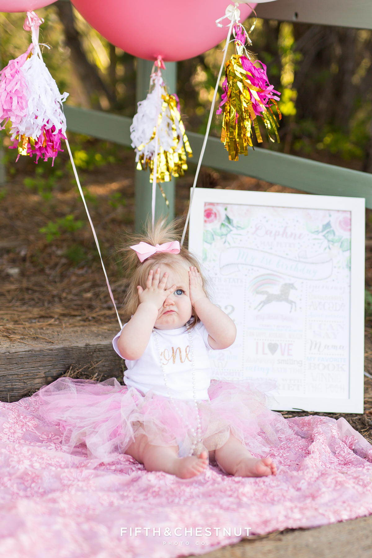 One year photos of baby girl making silly face at Kiva Beach with pink balloons and sign