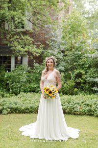 Bride stands in grass in front of farm house at the Twenty Mile house while holding a sunflower bouquet