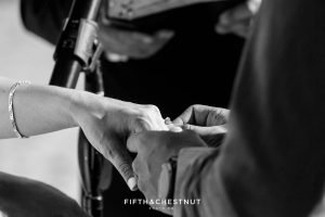 Black and white photo of the groom placing his wife's wedding ring on her ring finger