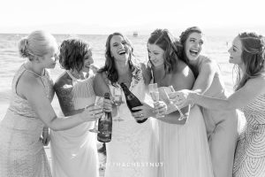 Bride and bridesmaids pop a bottle of champagne and laugh and smile for a Tahoe wedding on the beach of North Lake Tahoe