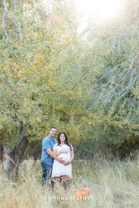 Husband and expecting wife stand in apple orchard with pumpkins for their fall maternity portraits