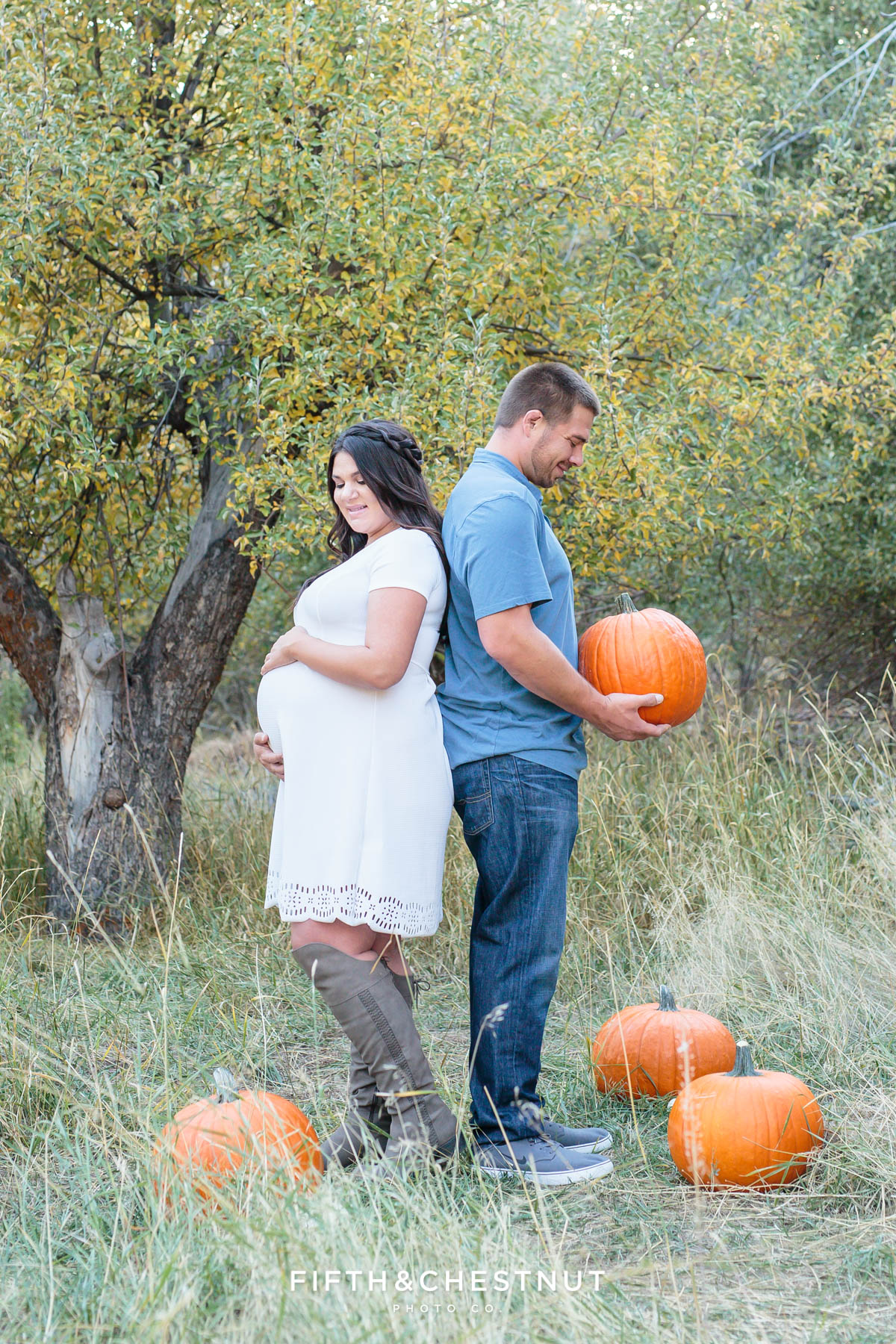 Close up of Husband and his pregnant wife stand back to back in an apple orchard surrounded by pumpkins while he looks down at a pumpkin he's holding and she looks down at her baby bump for their fall maternity portraits