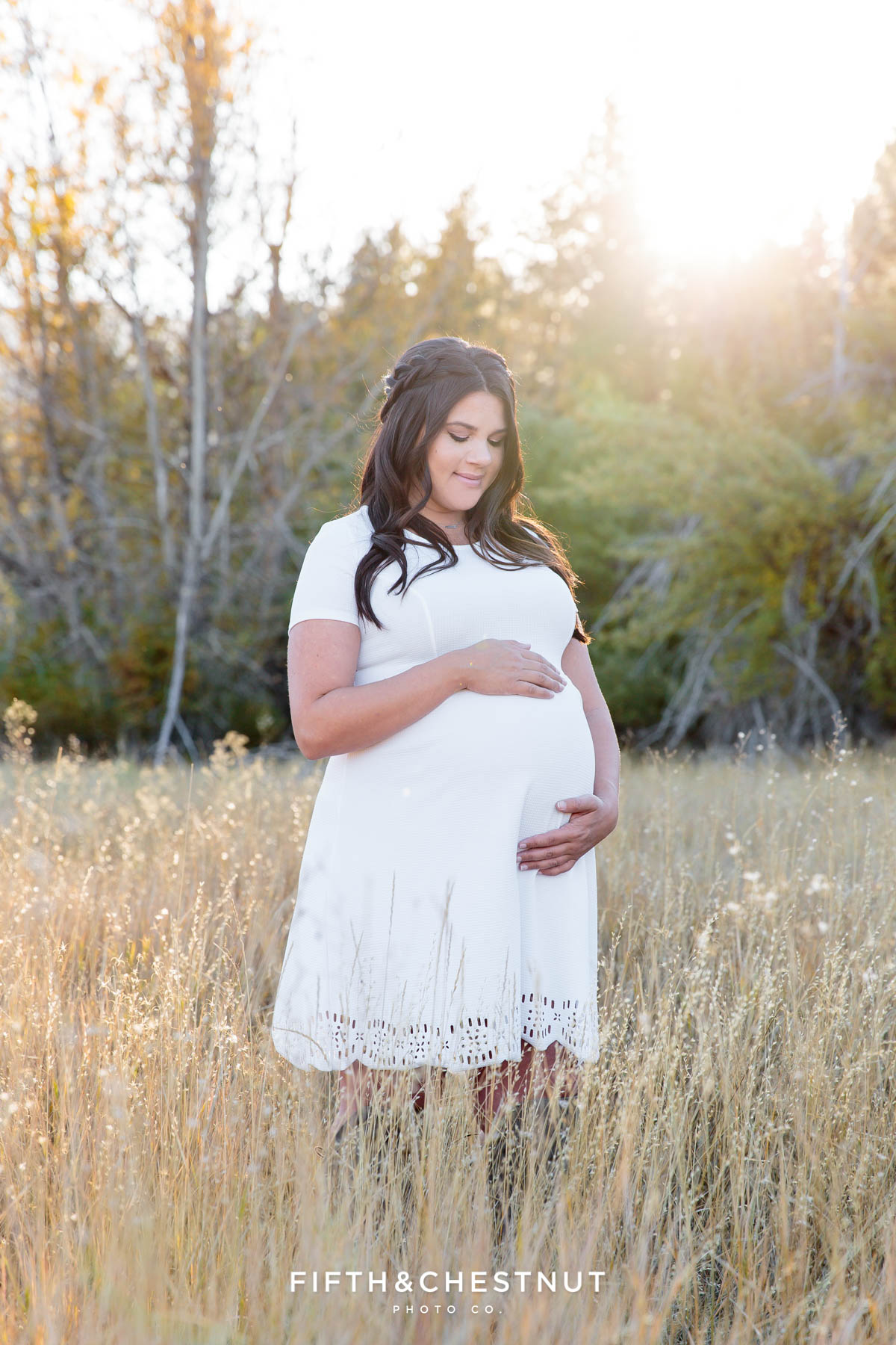 Pregnant woman in a white dress stands in a field and looks at her sweet baby bump for her fall maternity portraits