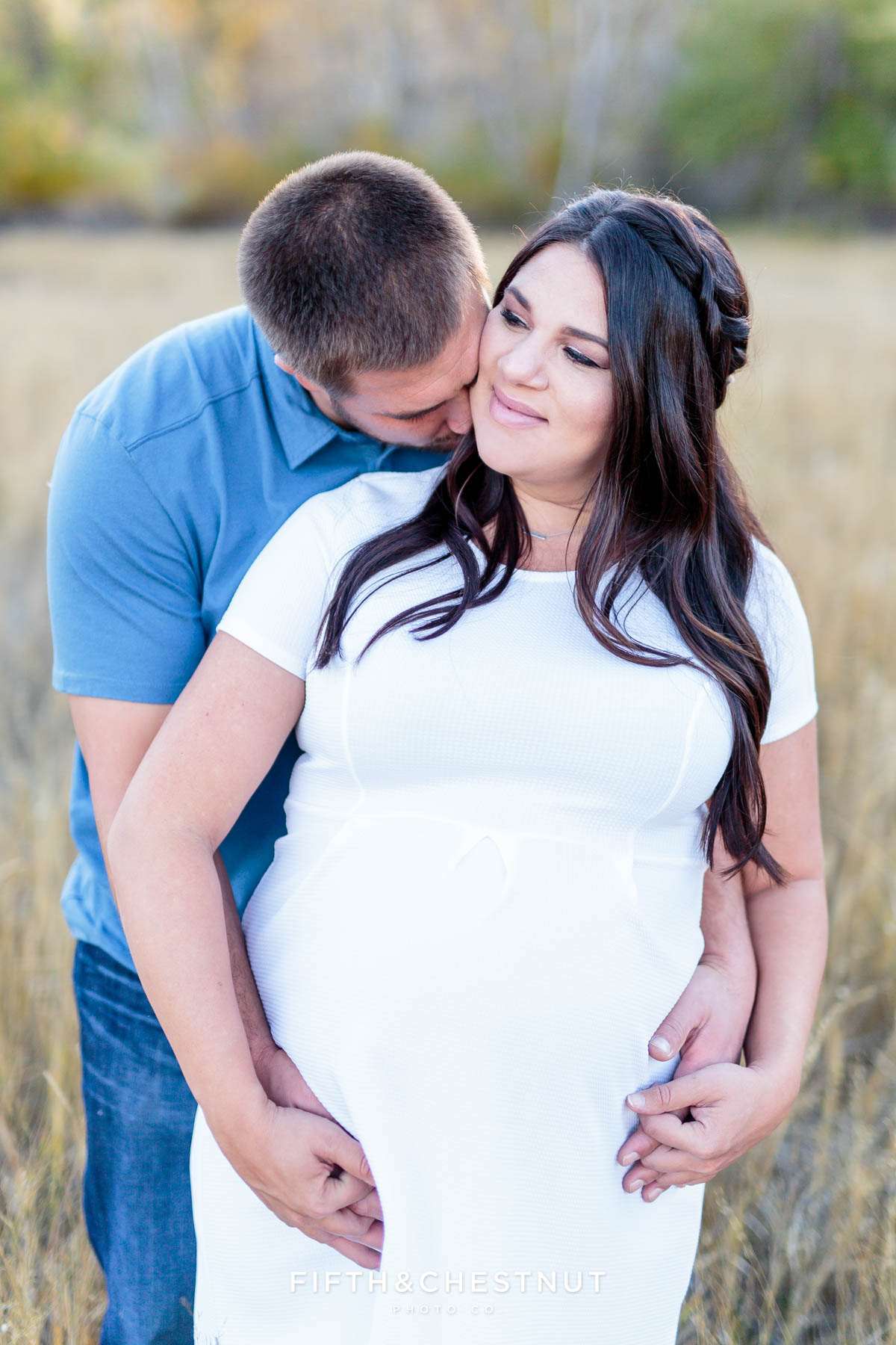 husband kisses his pregnant wife's neck as they hold hands in an open field for fall maternity portraits by Reno Maternity Photographer