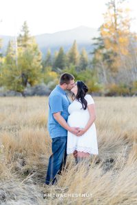 Expecting couple kiss in an open field with mountains and trees in the background for fall maternity portraits by Reno Maternity Photographer