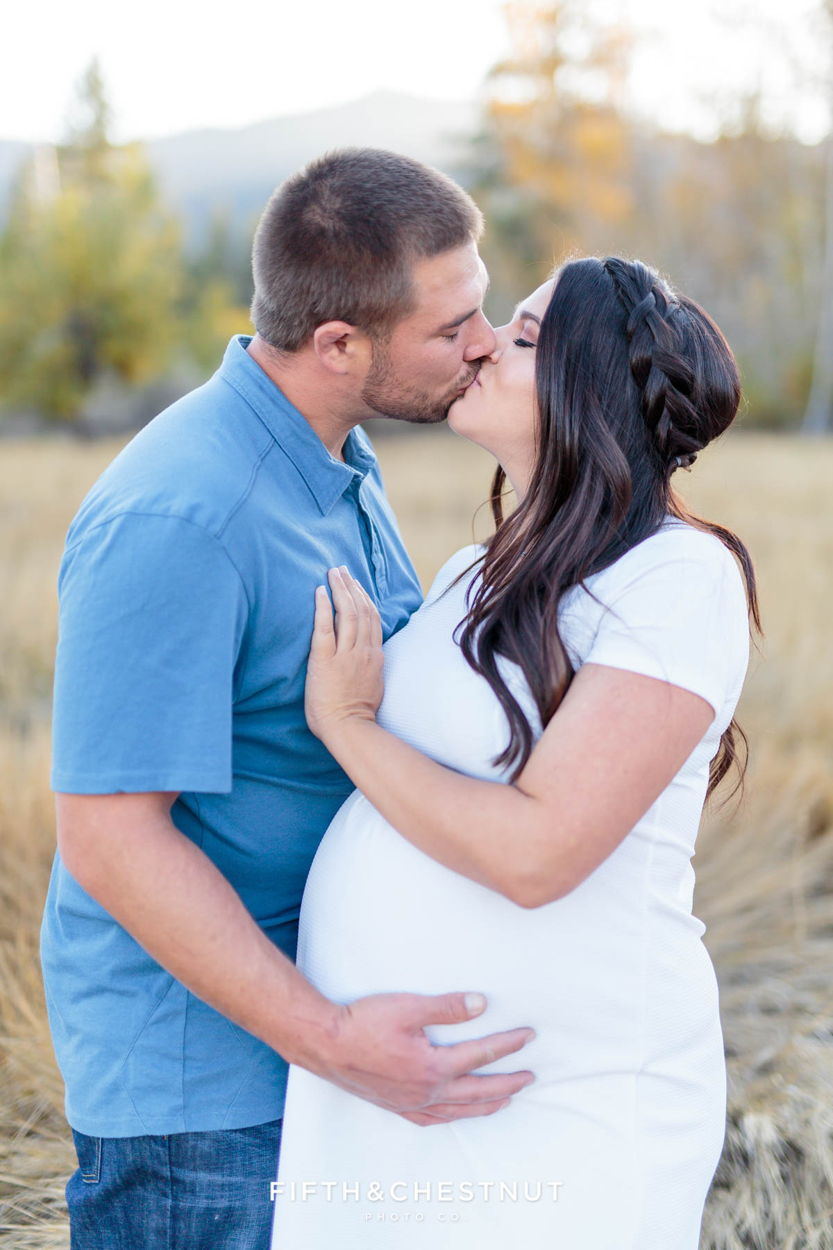 Husband and wife kiss for their fall maternity portraits by Reno Maternity Photographer in an apple orchard field with mountains behind them