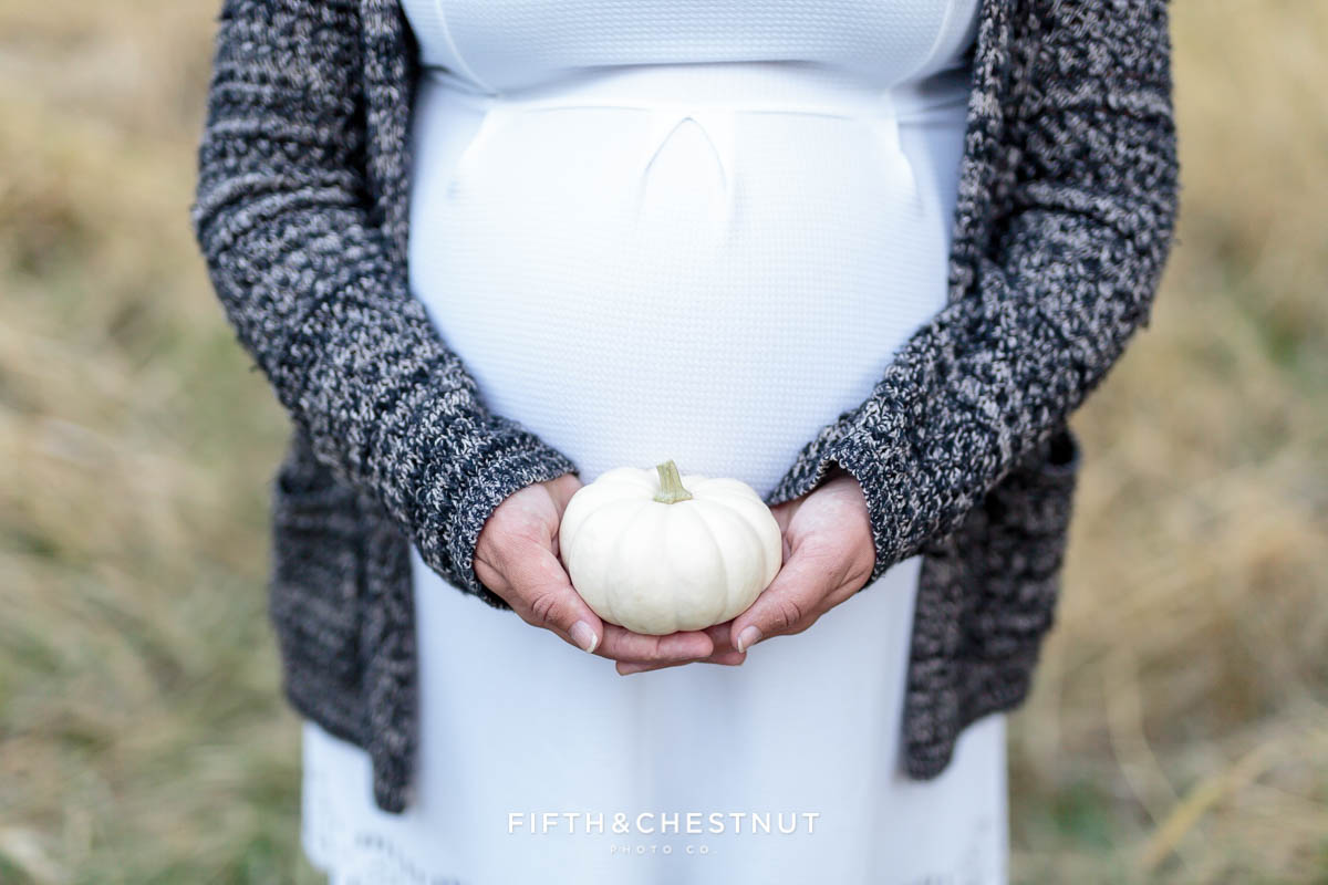 pregnant mama-to-be holds a small white pumpkin in front of her pregnant belly for fall maternity portraits by Reno Maternity Photographer