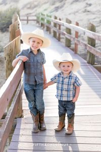 two little boys holding hands on a wooden path wearing plaid shits and cowboy hats