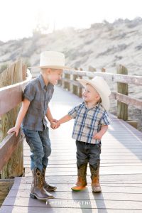 two little boys holding hands on a wooden path wearing plaid shits and cowboy hats giggling at each other