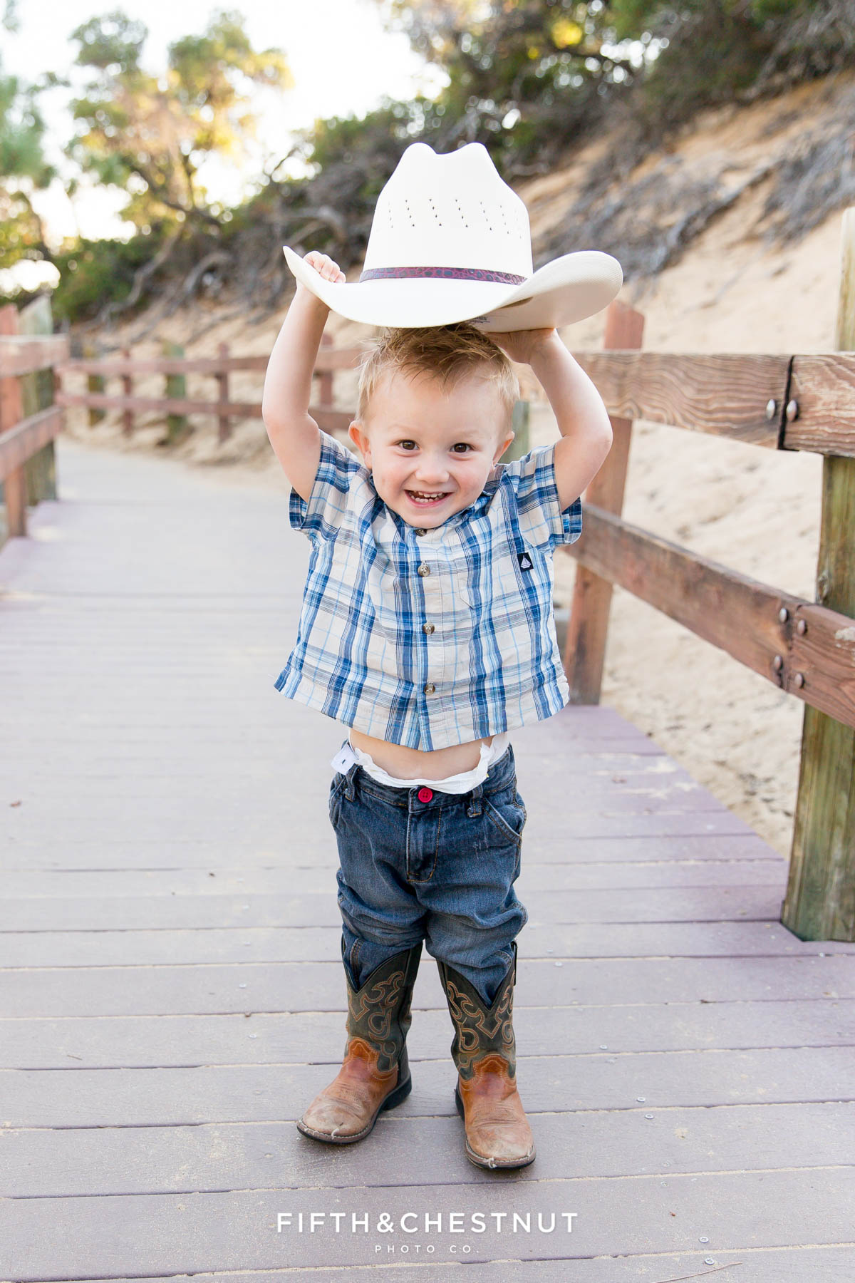Little boy holds cowboy hat above his head while wearing cowboy boots and a plaid shirt