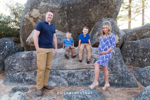 family wearing blue poses in front of big rocks at Sand Harbor for beautiful mama holds her pregnant belly for Tahoe Maternity Portraits