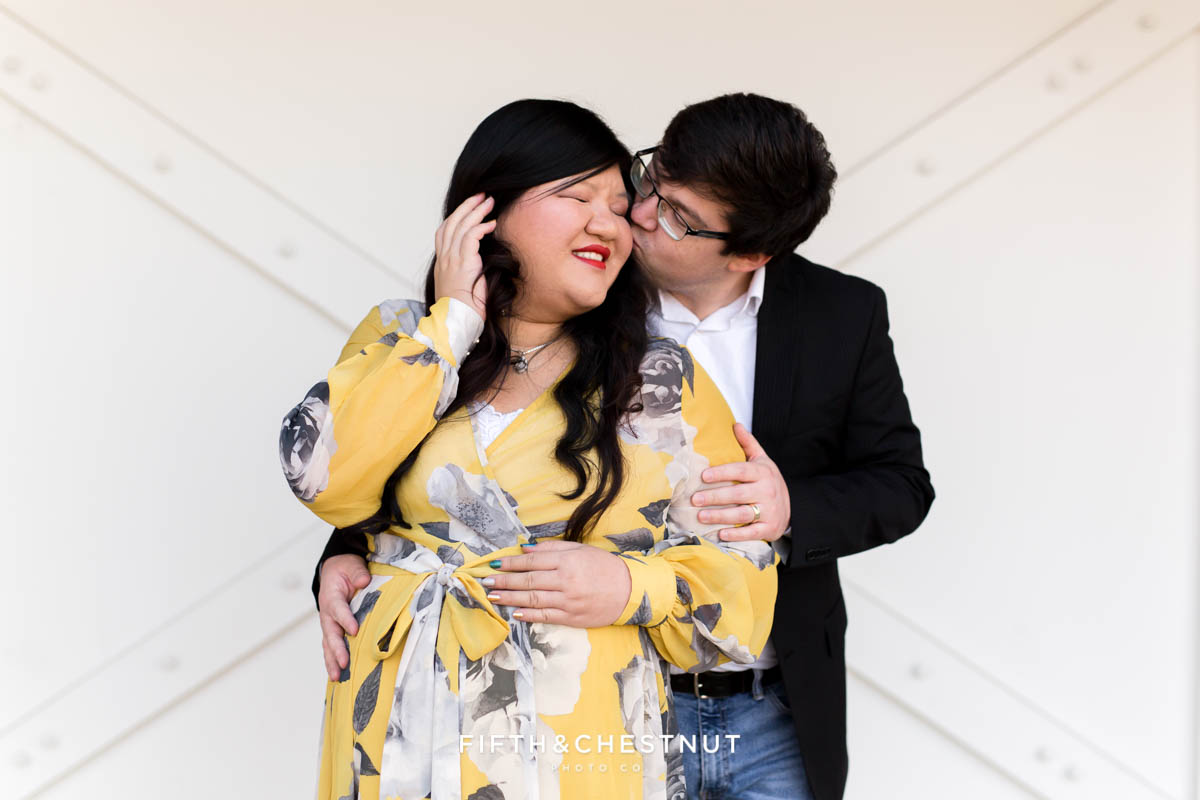 Husband kisses his pregnant wife while standing in front of a white barn door in Reno, NV for winter Reno maternity photos