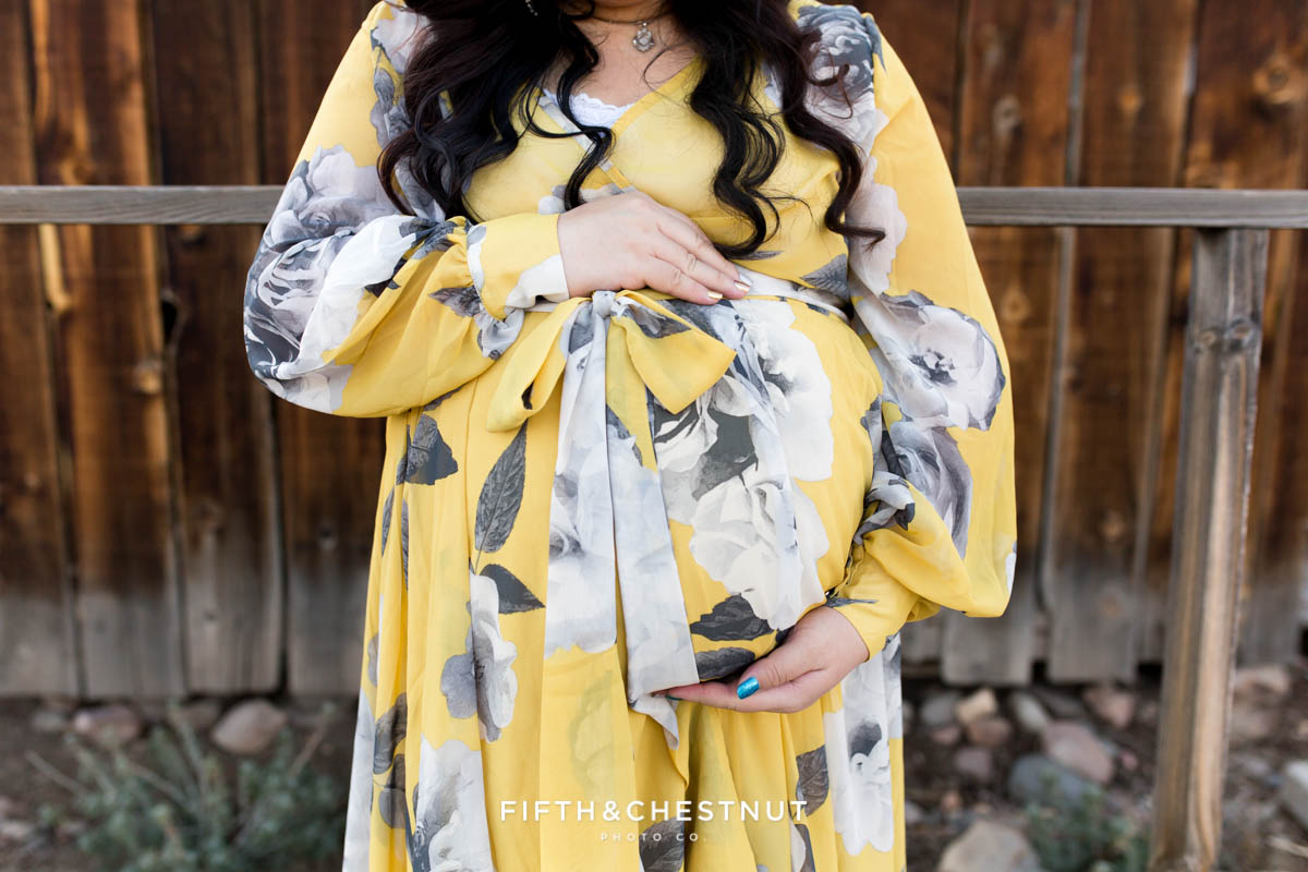 Pregnant woman in yellow and gray floral dress holds pregnant tummy while looking down surrounded by rustic western buildings in Reno, NV for maternity portraits by Reno Maternity Photographer