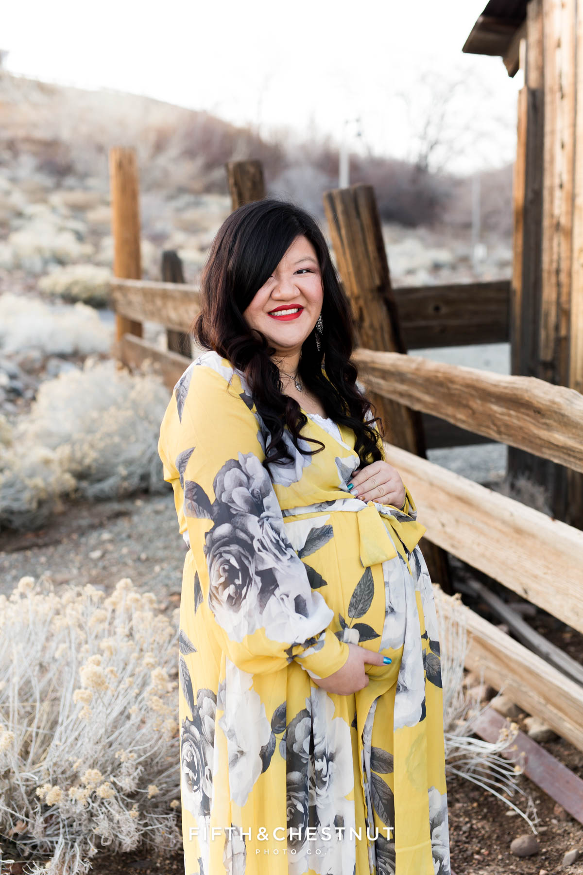 Pregnant woman in yellow and gray floral dress holds pregnant tummy while looking down surrounded by rustic fences and high desert foliage in Reno, NV for maternity portraits by Reno Maternity Photographer