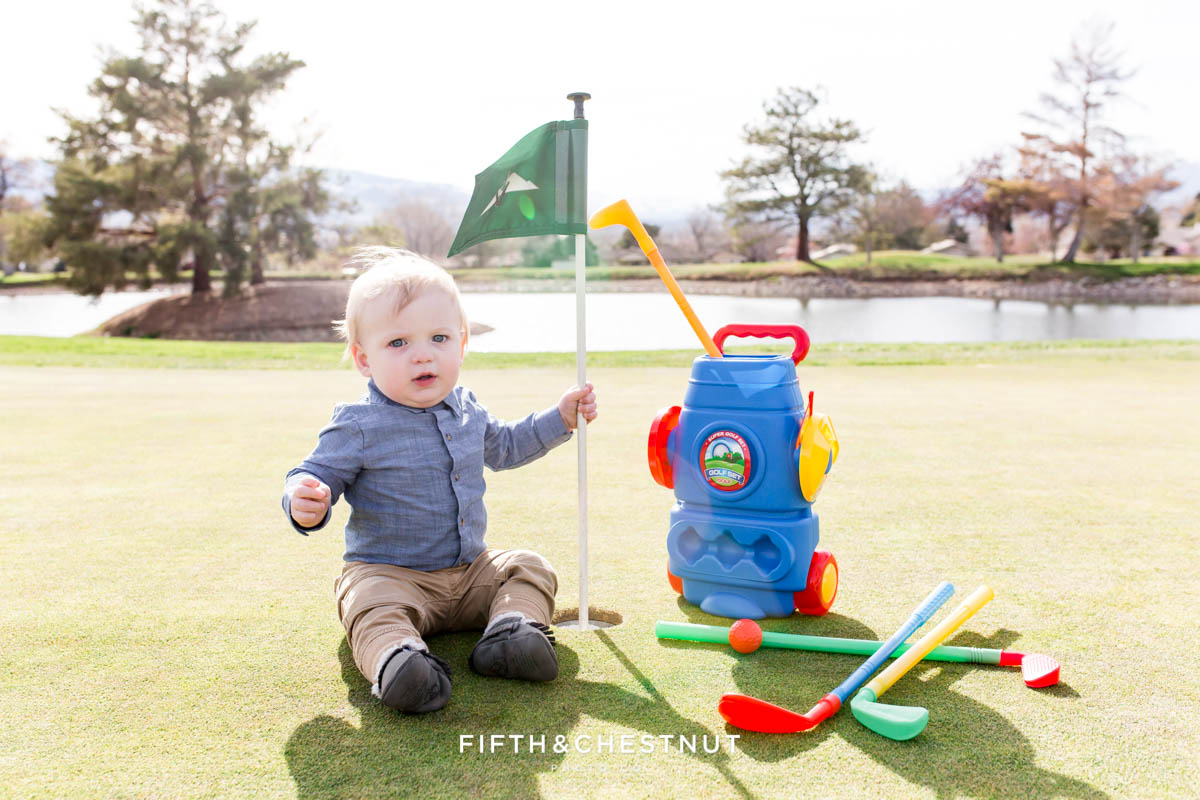 Baby boy holding flag on the green for his Golf Themed One Year Photos by Reno Baby Photographer surrounded by his toy golf clubs and golf balls on a sunny day