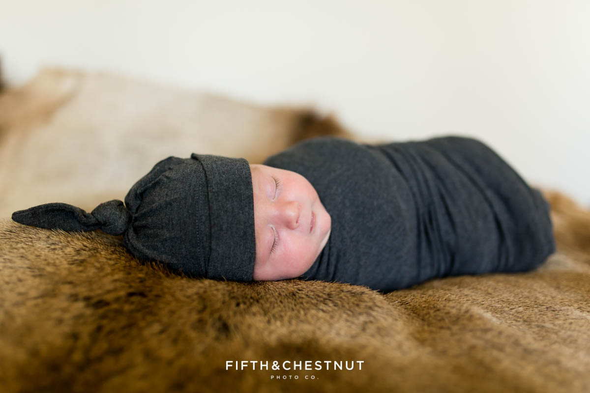 A baby boy wearing a gray swaddle and a newborn cap lays on an elk skin for his Reno newborn portraits by Reno Newborn Photographer