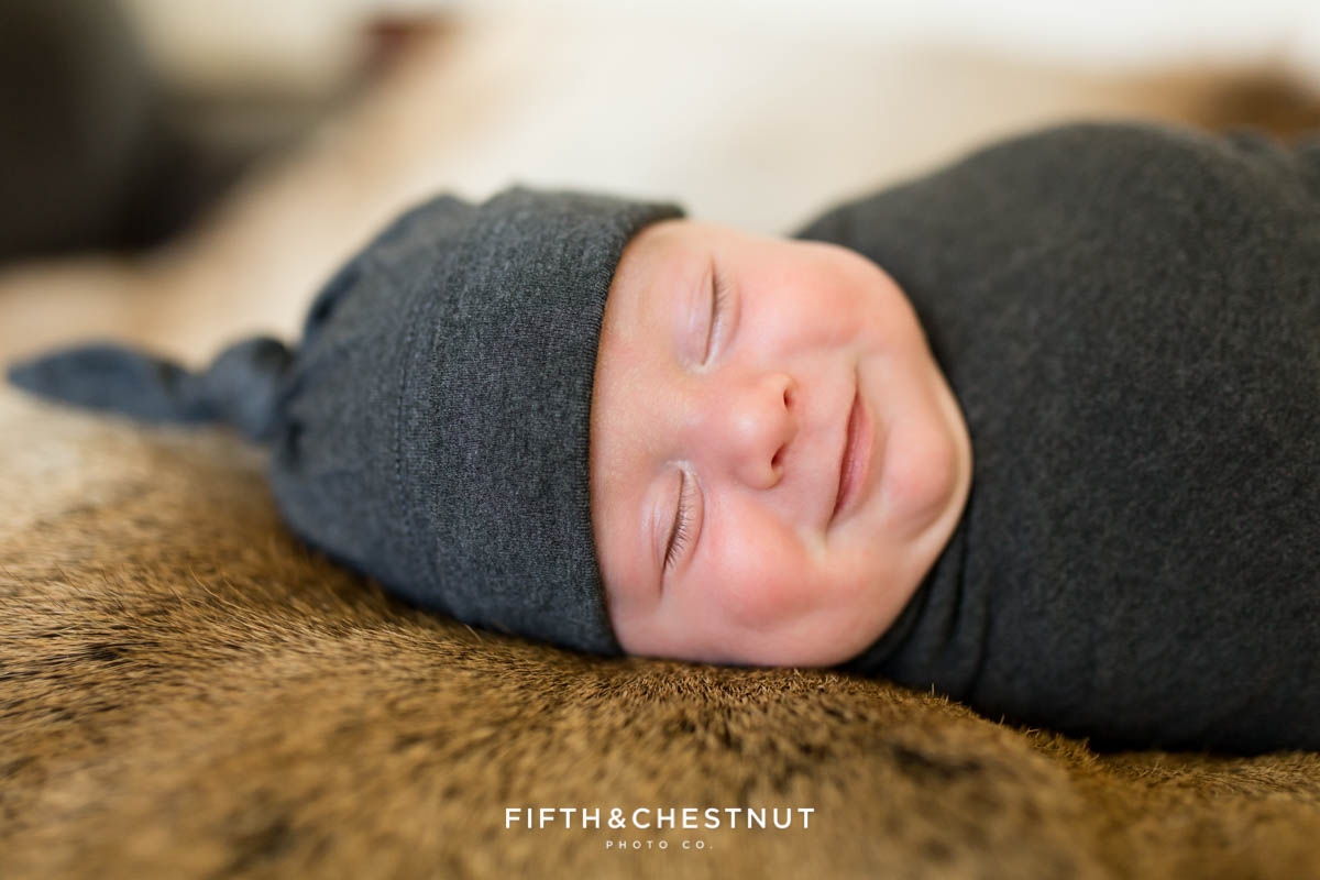 A smile caught on camera during a newborn photoshoot of a baby boy in a gray cap and gray swaddle on an elk skin pelt during his reno newborn photo session
