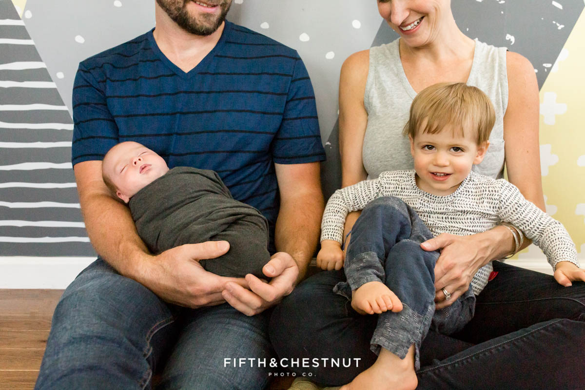 focus on a big brother and his new little brother while sitting in the laps of their parents while taking newborn baby photos
