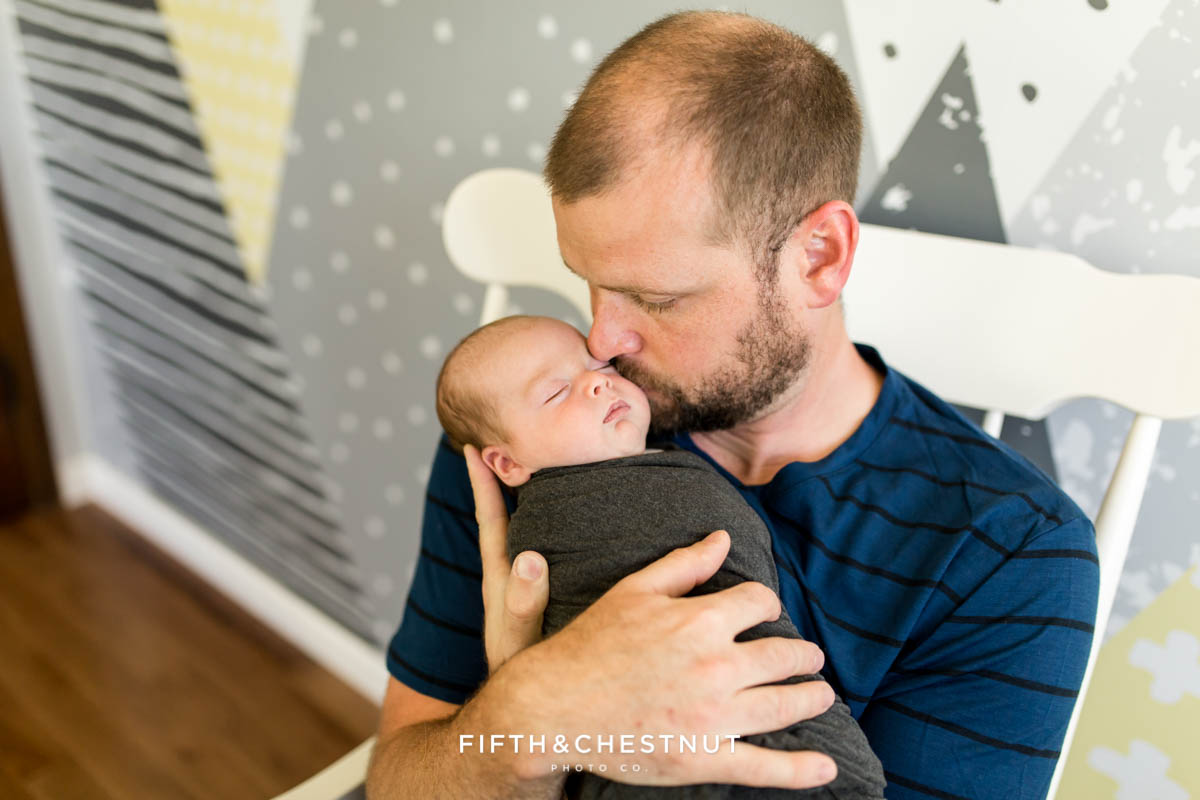 A dad kisses his newborn son on the cheek in the baby's gray and yellow nursery for Reno newborn photos