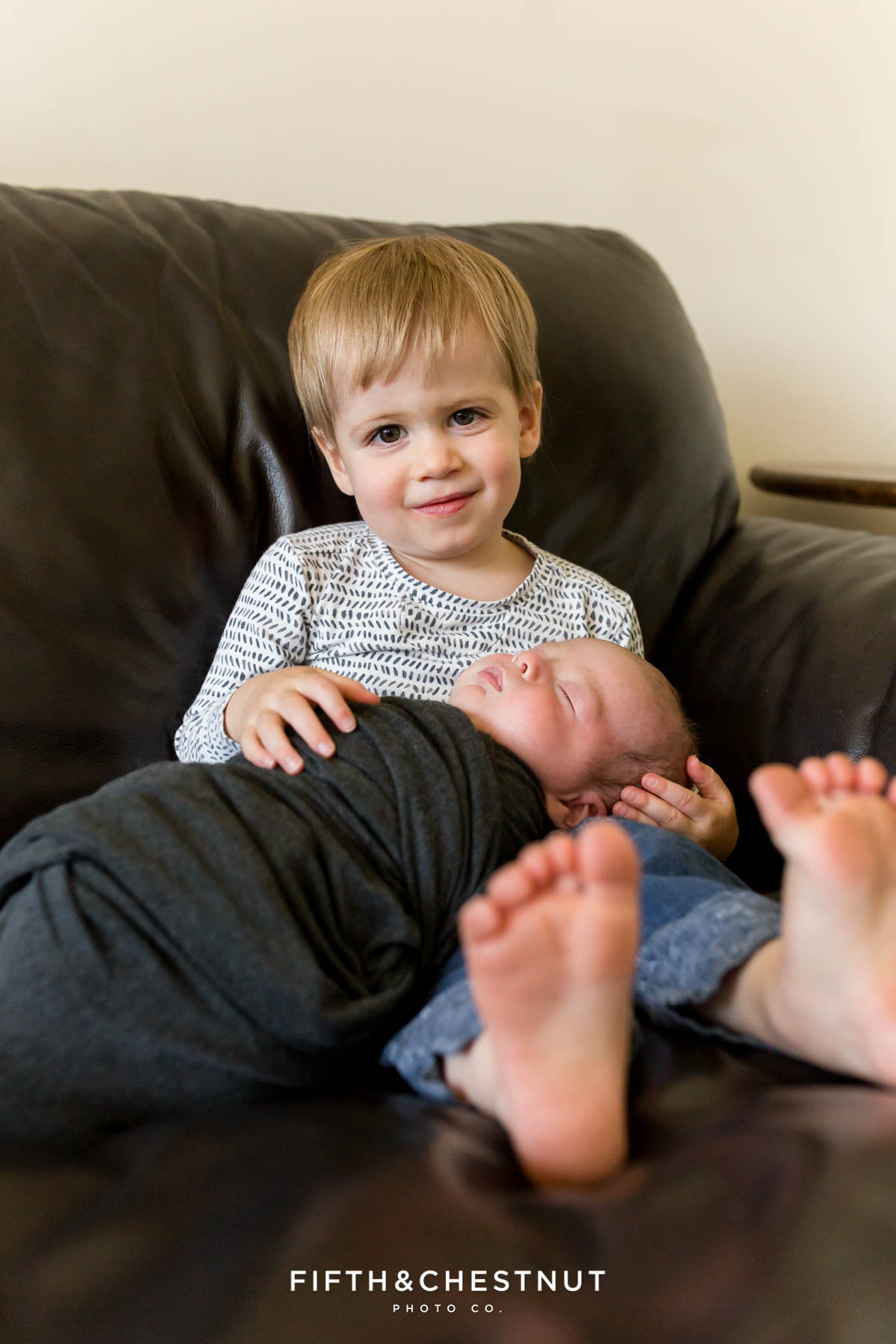 A color photo of a big brother toddler holding his new baby brother while sitting on a brown leather couch