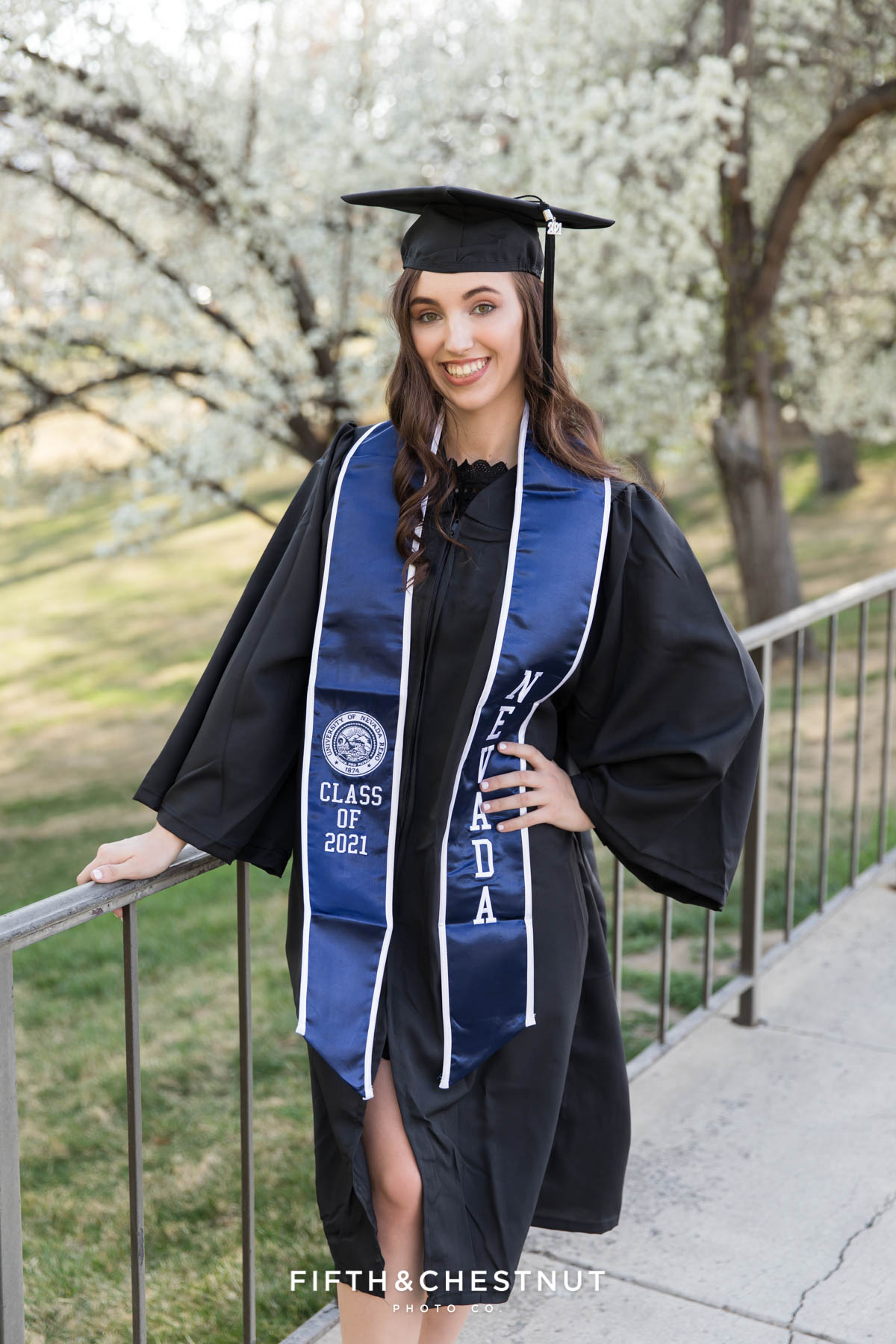 A beautfiul graduate in her cap and gown plus blue UNR sash in front of spring blossoms for her spring UNR graduation portraits