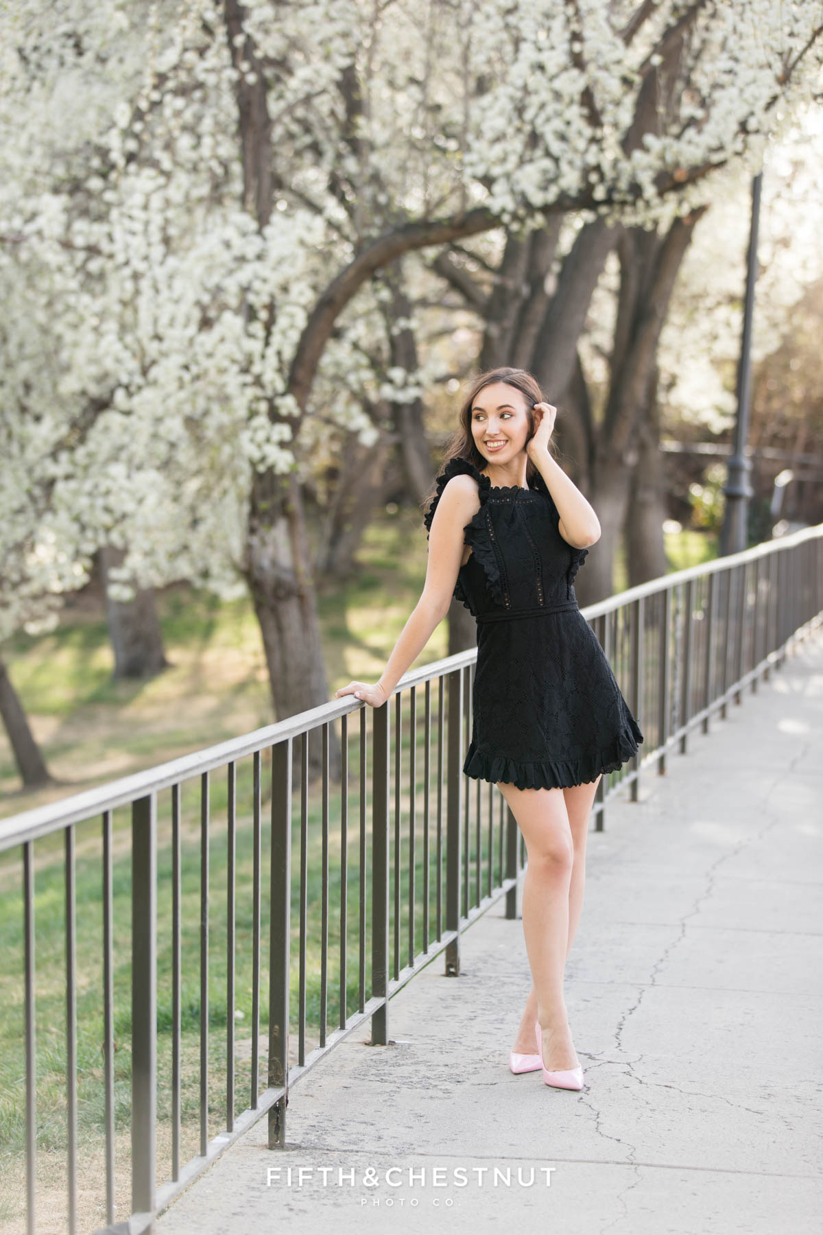 A UNR student leans on the fence of a walkway between the pond and the field of blossoming trees at UNR for her spring graduation pictures by Reno Graduation photographer